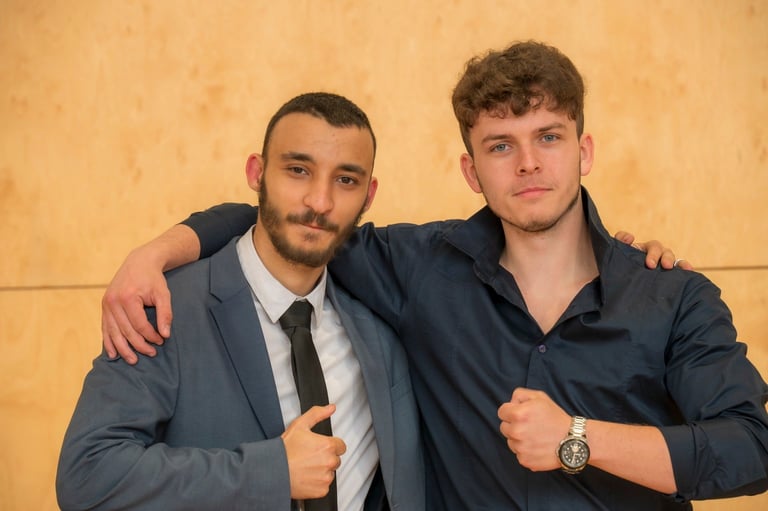 Two men posing together against a beige wall, one wearing a navy suit with black tie, the other in a dark navy shirt, both smiling at camera