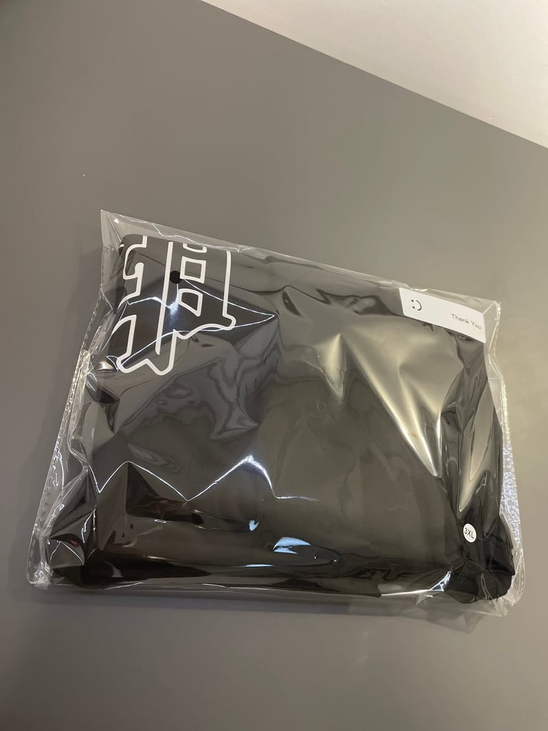 Black clothing item packaged in clear plastic wrap with white logo on gray wall