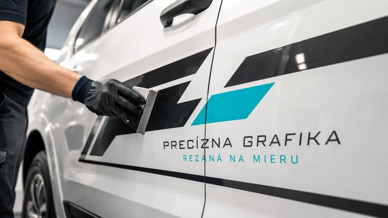 Person applying vinyl car wrap decal to white vehicle with Precizna Grafika company logo and turquoise design